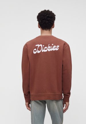 Person med kort krøllet hår vendt væk iført en brun sweatshirt med hvidt "Dickies" logo på ryggen og lyseblå jeans.
