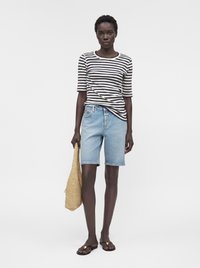 Femme portant une chemise rayée noire et blanche, un short en jean bleu clair, des sandales noires, tenant un sac tissé beige, debout devant un fond uni.