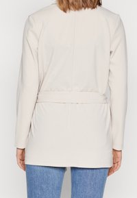 Vrouw in een lichtbeige jasje met riem en lange mouwen, gecombineerd met blauwe jeans, gezien van achteren tegen een effen achtergrond.