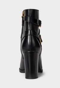 Botas de tornozelo em pele preta com um salto robusto, apresentando um fecho lateral, detalhes com tiras e uma textura suave.