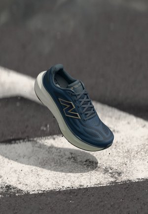 Zapatilla de running New Balance azul suspendida sobre una carretera asfaltada con marcas blancas pintadas y gravilla esparcida.