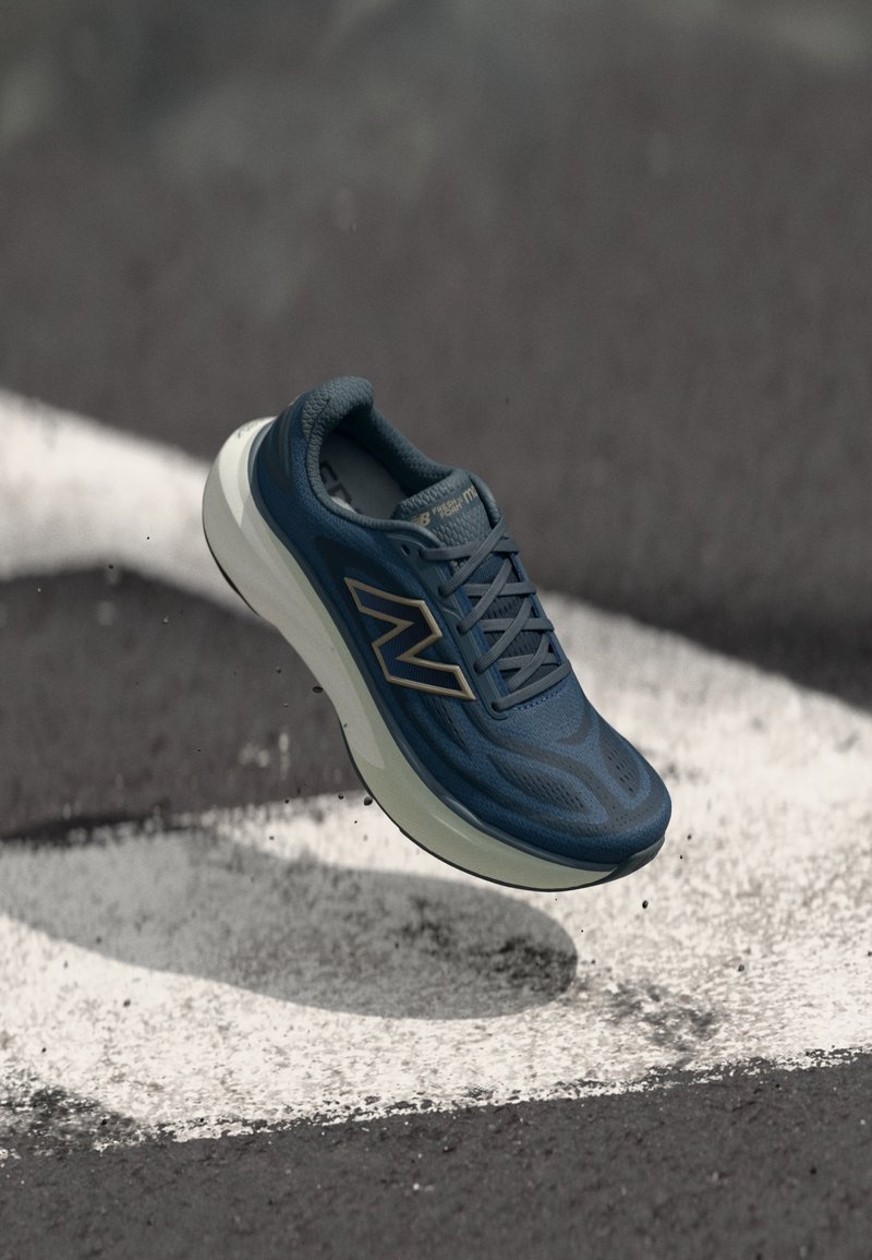 Μπλε δρομικό παπούτσι New Balance, αιωρούμενο πάνω από έναν ασφαλτοστρωμένο δρόμο με λευκές βαμμένες γραμμές και διάσπαρτα χαλίκια.