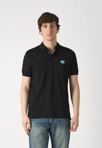Svart polo-shirt laget av bomull, med klassisk krage, korte ermer, og en lys blå logo-patch på venstre bryst. Brukt sammen med blå jeans.