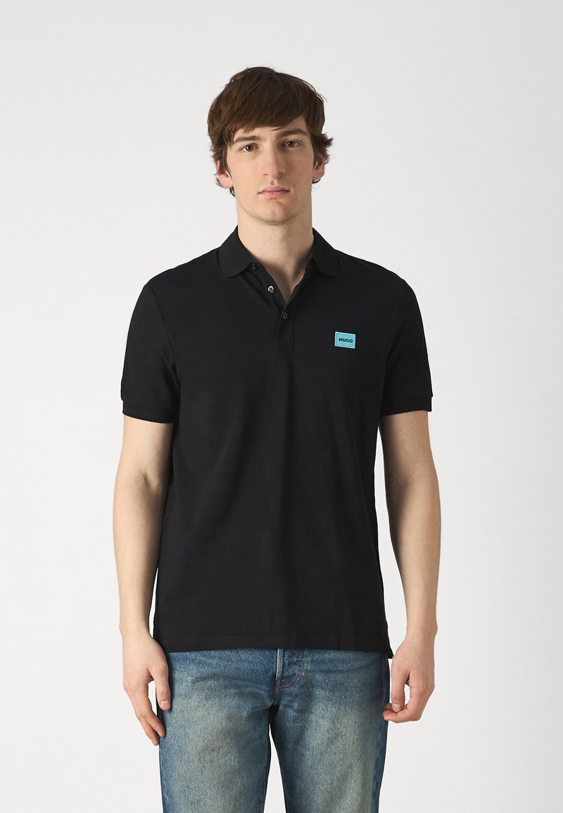 Svart polo-shirt laget av bomull, med klassisk krage, korte ermer, og en lys blå logo-patch på venstre bryst. Brukt sammen med blå jeans.