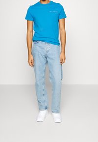 T-shirt in cotone blu con collo a giro e logo "HILFIGER", abbinata a jeans in denim blu chiaro e sneakers bianche. Look semplice e casual.
