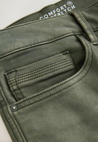 Oliivrohelised denim püksid, millel on sile tekstuur, lametaskud ja nähtavad õmblemise üksikasjad. Sildil on kirjas "Comfort Stretch".