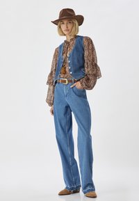 Gilet in denim blu con bottoni argentati, blusa marrone a fantasia, jeans larghi blu, cintura marrone e cappello da cowboy marrone con una fascia.