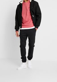 Veste noire avec une capuche en fourrure, sweat à capuche rouge et joggers noirs. Des baskets blanches complètent la tenue, ajoutant un look décontracté.