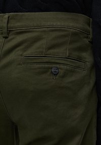 Gros plan sur la poche arrière d'un pantalon vert olive avec un bouton noir, montrant la passants de ceinture et un fond en tissu sombre.