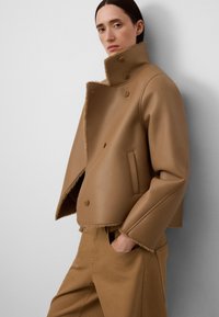 Hellbraune, taillierte Jacke aus glattem Leder mit einem hohen Kragen, doppelten Knöpfen und fransigem Saum. Kombiniert mit passenden hellbraunen Hosen.
