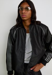 Chaqueta bomber negra con acabado brillante, rayas grises en contraste en las mangas, cuello y puños de punto. Llevada con una camiseta sin mangas blanca y gafas de sol.