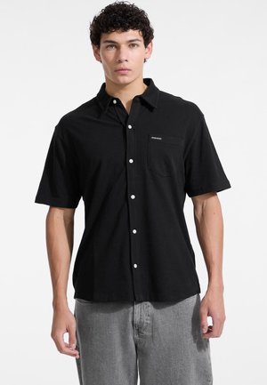 Black short-sleeve button-up shirt in soft fabric, featuring a chest pocket and white buttons, paired with light gray pants.