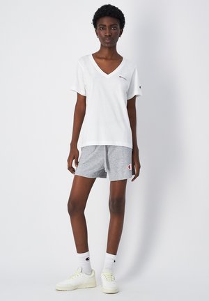 Weißes V-Ausschnitt-T-Shirt, kurze graue Baumwollshorts mit Logo-Patch, weiße Socken und hellgelbe Sneakers. Einfaches Design, lockere Passform.