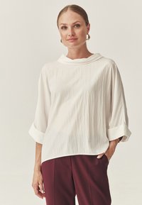 Vrouw met haar opgestoken, draagt gouden oorbellen, een losse witte blouse met opgerolde mouwen en bordeauxrode broek, staande met één hand in de zak.