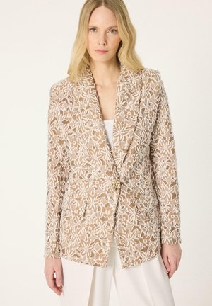 Donna con blazer beige con sovrapposizione in pizzo floreale bianco e bottone dorato, abbinato a pantaloni bianchi, di fronte a uno sfondo neutro.