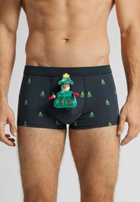Černé boxerky s opakujícím se vzorem zelených vánočních stromků. Uprostřed je umístěn plyšový tučňák v zeleném oblečení a klobouku.