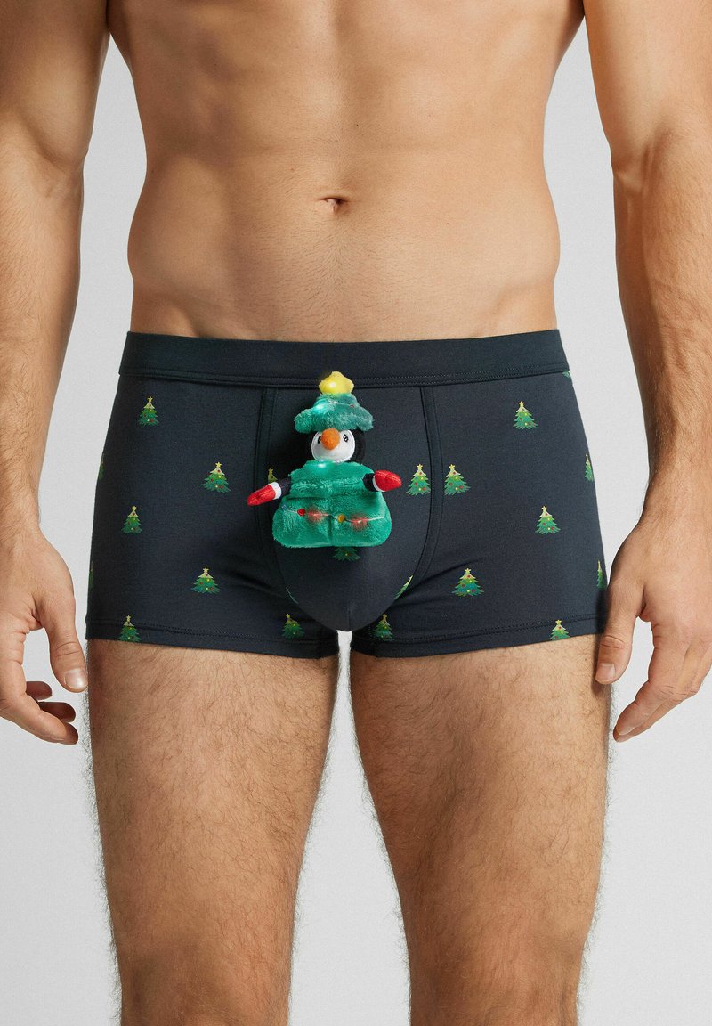 Černé boxerky s opakujícím se vzorem zelených vánočních stromků. Uprostřed je umístěn plyšový tučňák v zeleném oblečení a klobouku.