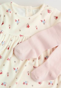 Virágmintás krémszínű ruha rózsaszín harisnyával. A ruha apró piros és kék virágokkal, kerek nyakkal és redőzött részletekkel rendelkezik. A harisnya puha rózsaszínű.