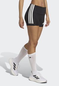 Czarne szorty sportowe z trzema białymi pionowymi paskami po bokach, w połączeniu z białymi skarpetami do kolan i białymi butami z czarnymi akcentami.
