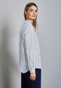 Randig skjorta med knappar i vitt och marinblått, med krage, långa ärmar och avslappnad passform, kombinerad med mörkblå jeans.