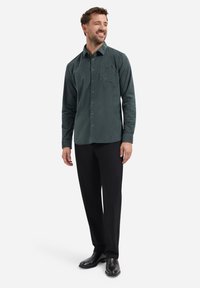 Donkergroen overhemd met knopen en een zak, gedragen met zwarte broek en laarzen. De stof lijkt gestructureerd en licht van gewicht.