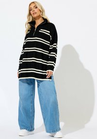 Svart- och vitrandig overdimensional stickad tröja med dragkedja vid kragen, tillsammans med vida ljusblå jeans och vita sneakers.