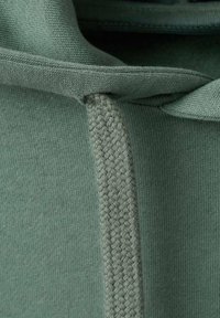 Tela de algodón verde con una textura suave, que presenta un detalle de cordón trenzado a lo largo del borde y costuras finamente realizadas.