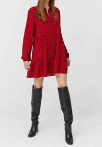 Robe fleurie rouge avec un devant boutonné, des manches longues et un ourlet volanté. Portée avec des bottes en cuir noires montantes. Texture lisse.