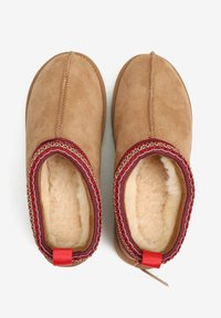 Jasnobrązowe kapcie zamszowe z miękką podszyciem z polaru, czerwono-srebrne nici ozdobne na kołnierzu oraz mała czerwona pętelka z tyłu.
