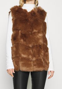 Kvinna som bär en ärmlös brun fuskpälsväst över en vit långärmad topp och svarta byxor, visad från axlarna till höfterna.