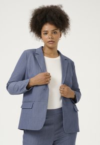 Blazer blu, con texture, colletto a revers, un bottone e due tasche frontali, indossato su un top bianco, con una leggera vestibilità sartoriale.