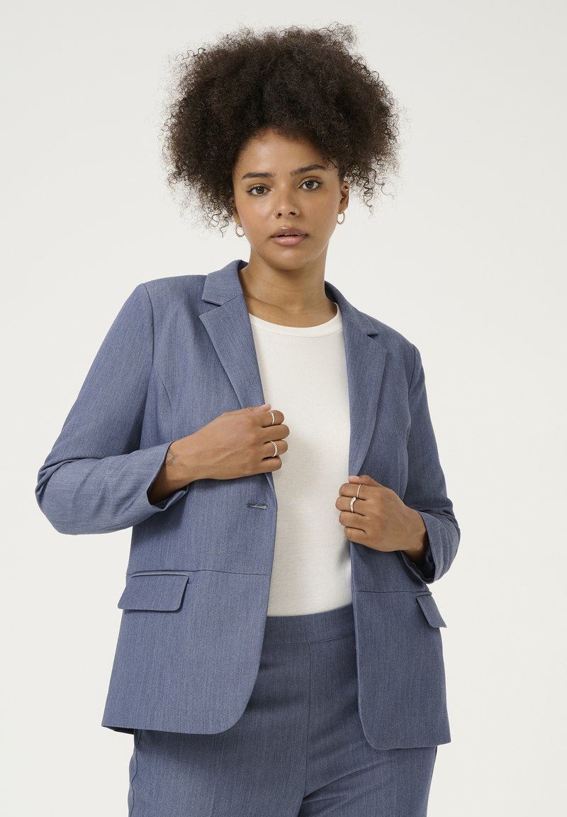 Blazer blu, con texture, colletto a revers, un bottone e due tasche frontali, indossato su un top bianco, con una leggera vestibilità sartoriale.