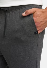 Nærbilde av en hånd som hviler i frontlommen på mørkegrå, teksturerte joggebukser med svart snor og liten logolapp.