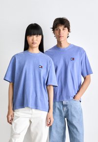 Divi jaunieši, kuri valkā gaiši zilo Tommy Jeans t-kreklu, viens saskaņots ar baltām saplēstām biksēm, bet otrs - ar gaiši zilām džinsām, stāv blakus viens otram.