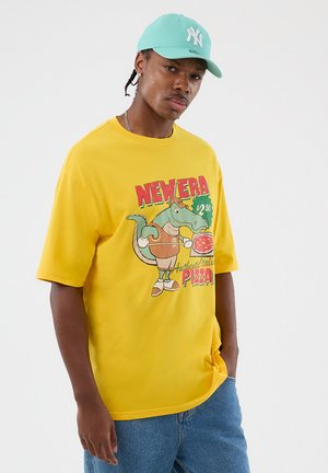 Νεαρός άνδρας με τιρκουάζ καπέλο, κίτρινο μπλουζάκι με καρτούν αλιγάτορα που κρατά πίτσα και μπλε τζιν, που στέκεται μπροστά σε απλό λευκό φόντο.