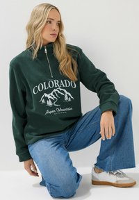 Pullover in pile verde con mezza zip, caratterizzato da una grafica di montagne bianche e il testo "COLORADO". Abbinato a jeans blu a gamba larga e scarpe da ginnastica argentate.