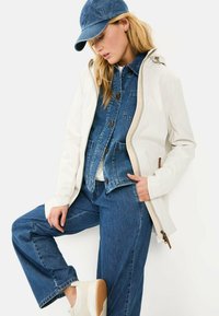 Witte waterdichte jas met beige ritssluiting, gelaagd over een blauwe denimjas en blauwe wijde jeans. Inclusief een denim pet en witte sneakers.