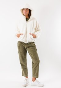 Weißer Fleece-Zip-Hoodie mit Kapuze und Fronttaschen, kombiniert mit olivgrünen Cordhosen und weißen Sneakern. Sichtbare strukturierte Oberflächen.