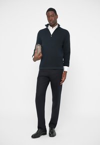 Tmavomodrý sveter s textúrou a zipsovým golierom, pod ním biela košeľa, doplnené o tmavé spoločenské nohavice a čierne topánky. V ruke drží sivý doplnok.