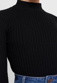 Gros plan sur un torse portant un haut noir côtelé à manches longues avec col montant, rentré dans un jean en denim bleu taille haute.