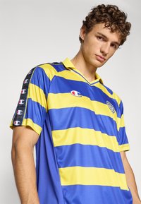 Ung mand med krøllet hår iført en blå og gul stribet sports T-shirt med Champion-logoer på brystet og ærmerne.