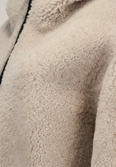 Tessuto teddy fleece beige con superficie morbida, testurizzata e soffice, e un bordo della cerniera nera visibile.