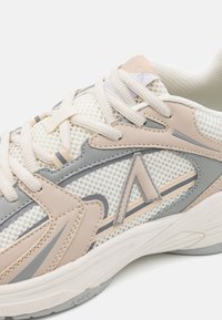 Leichter Sneaker aus Mesh-Material mit beigen und grauen Akzenten, strukturierten Oberflächen und kontrastierender Naht entlang der Seiten.