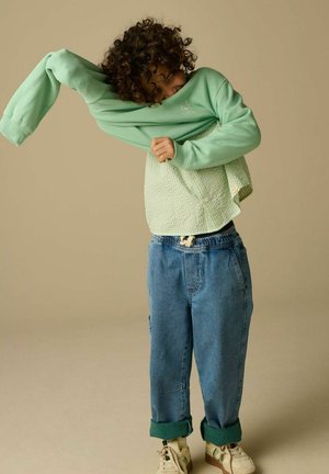 Enfant aux cheveux bouclés tirant un sweat-shirt vert par-dessus la tête, portant un jean bleu ample et des baskets beige sur un fond beige uni.