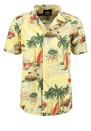 Mand iført en gul tropisk print kortærmet skjorte, sorte op-rullede jeans, brune sko, der holder en marineblå hat i den ene hånd.