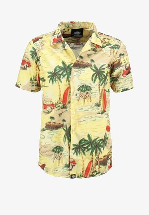 Man draagt een geel tropisch print shirt met korte mouwen, zwarte opgerolde spijkerbroek, bruine schoenen, houdt een marineblauwe hoed in één hand.