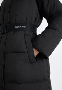 Melnā pūkainā jaka ar jostu vidukļa daļā, ar spīdīgu audumu, quilted struktūru un divām sānu kabatām. Jostā ir "Calvin Klein" logo.