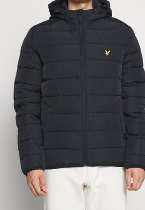 Personne portant une doudoune noire à capuche avec des coutures horizontales et un petit logo d’oiseau jaune sur la poitrine, associée à un pantalon blanc.