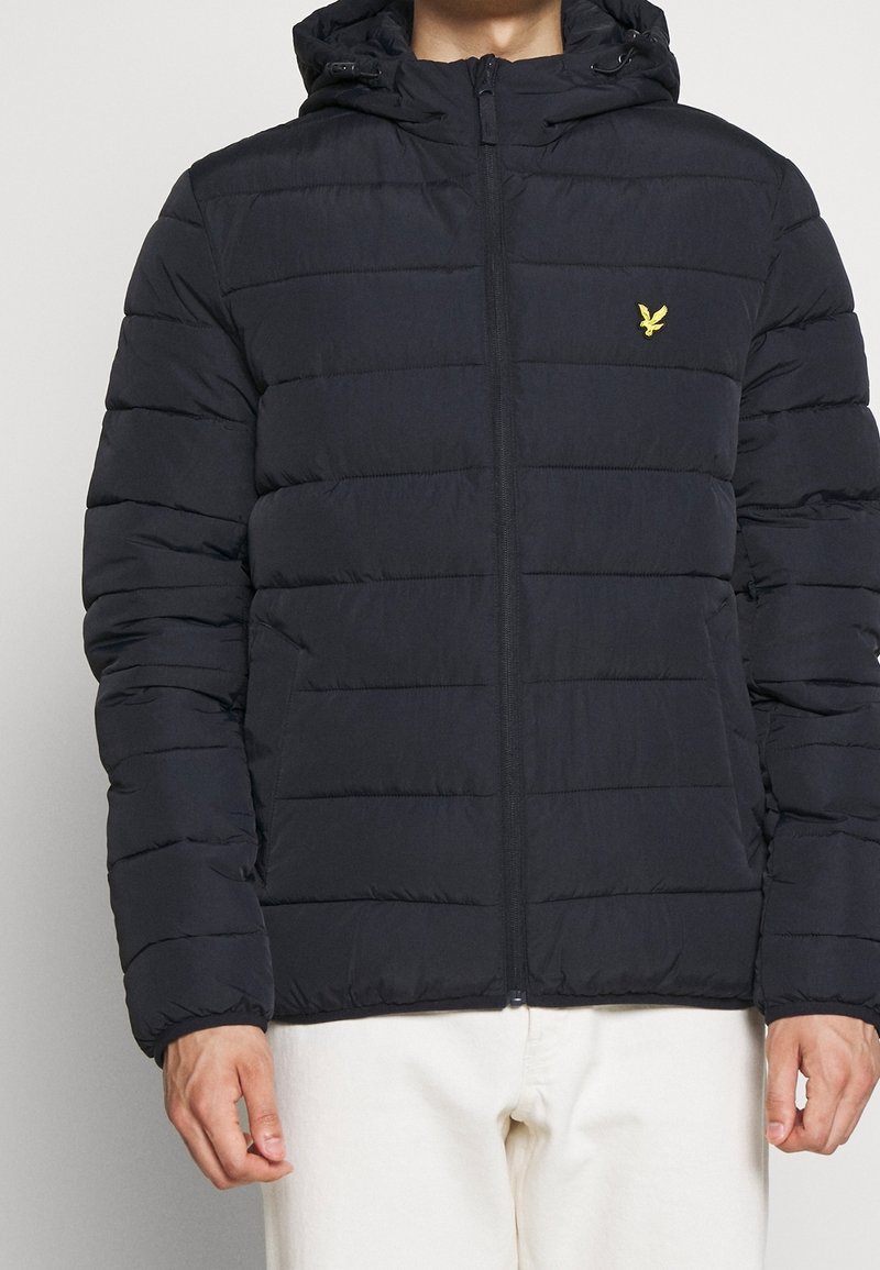 Personne portant une doudoune noire à capuche avec des coutures horizontales et un petit logo d’oiseau jaune sur la poitrine, associée à un pantalon blanc.
