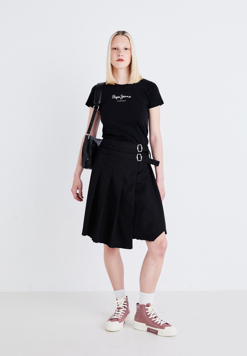 Camiseta negra con logotipo blanco, combinada con una falda plisada negra con cinturones de doble hebilla, y zapatillas altas rosas con suelas blancas.